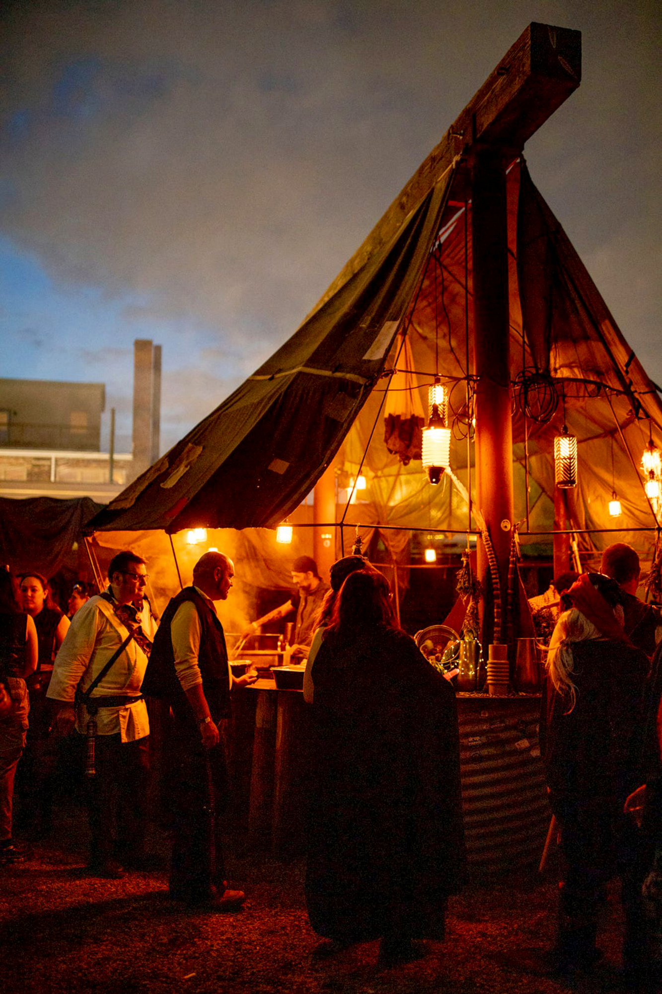 Customers form a line around a vendor stall, made to look like a make-shift hut. Inside, pots of food steam and workers portion out meals. Flickering lanterns hang above, and the countertop is adorned with alien-looking displays of unusual produce.