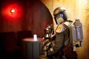 An adult and child, both dressed in Mandalorian style armor, look off-camera to the left while standing in familiar arched alcoves inspired by the Mos Eisley cantina from "Star Wars"