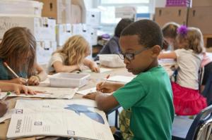 Elementary school students working on an art project at a classroom table, with a young boy in glasses and a green shirt painting in the foreground.