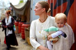 A woman dressed in white cradles a baby in a white fleece smock, and the baby holds a plush of the Mandalorian character "Grogu." Robed patrons pass in the outdoor marketplace.