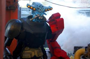 A blue, insect-headed alien creature with a red robotic arm stands in an industrial looking environment as smoke billows in the background