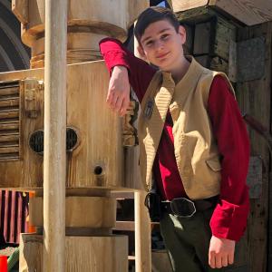A preteen boy dressed in a red shirt and tan vest poses next to a futuristic looking prop inspired by the moisture vaporators from Star Wars