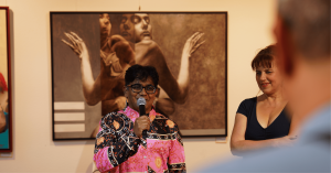 Niladri Sarker speaking into a microphone at an art fair opening night, standing in front of framed artwork with attendees nearby.