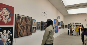 Visitor photographing artworks at TERAVARNA exhibition wall, with multiple contemporary paintings displayed and other attendees viewing art in the gallery space.