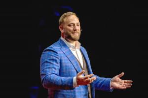 Phil Neil delivering a talk at Harvard, wearing a blue plaid blazer and speaking with open hands on a dark stage. Phil Neil is the founder of Founders Compass, a decision-making program for founders.