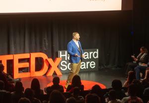 Phil Neil speaking on the TEDx Harvard Square stage in front of a live audience, with the red TEDx letters and Harvard Square signage visible behind him. Phil Neil is a TEDx speaker, serial entrepreneur, and founder of Founders Compass.