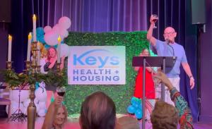 Scott Pridgen, Executive Director of Keys Health and Housing, raises a glass in a toast at a community celebration marking the organization's new name and 40th anniversary. The Keys Health and Housing logo is visible on a backdrop behind him.