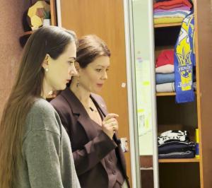 Two women stand inside a recreated Ukrainian child's bedroom, looking at a wardrobe stocked with folded clothes and Ukrainian football scarves. The moment captures Nobel Peace Prize laureate Oleksandra Matviichuk visiting the Empty Beds installation.