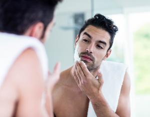 A man applying shaving cream to his face while looking in a bathroom mirror, preparing to shave