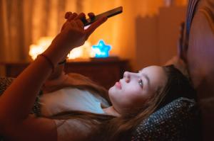 A teenage girl lies in a dark bedroom, her face illuminated by the glow of her smartphone.