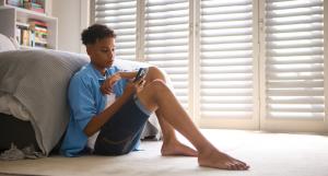 A teenage boy sits alone on the floor of his bedroom, focused on his smartphone.