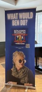 Blue Image banner of What Would Ben Do stand up book sale sign (photo credit: PLA Media). Banner includes book cover of Benjamin Franklin: America’s First Rockstar by Pamela Lewis