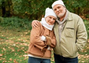A smiling older couple embraces outdoors in autumn, leaning close together against a backdrop of green and gold foliage.