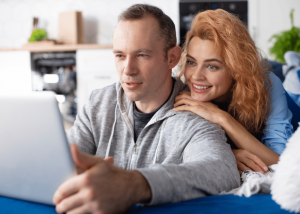 A smiling couple sits together on a couch, leaning close as they look at a laptop screen together in a cozy home setting.