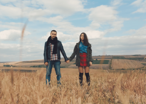 A young couple stands hand in hand in an open field under a cloudy sky, facing forward with serious expressions and a slight physical distance between them.