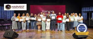 Group photo of Harvest Community School staff and management holding certificates with IBCCES CEO, Myron Pincomb onstage, with school and accreditation logos in the background.