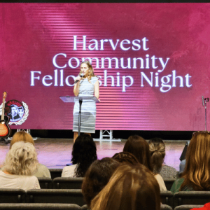 A representative from Harvest Community School addresses the audience during Harvest Community Fellowship Night in a dimly lit room with a large pink screen.