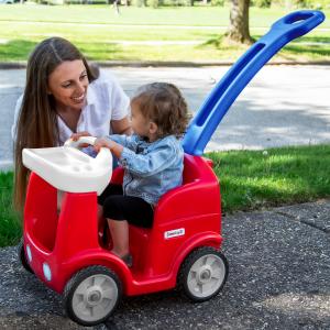 A mom kneeling next to her daughter as she sits in a red toy car.
