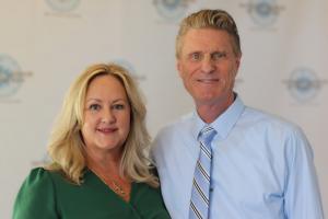 A smiling lady in green dress and gentleman in shirt and tie, on a step and repeat carpet event.