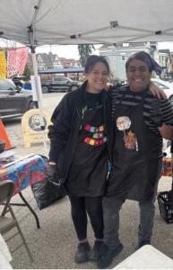 Owners of Lula La Chula stand together smiling under a canopy at an outdoor farmers market booth, wearing aprons as they prepare and serve food