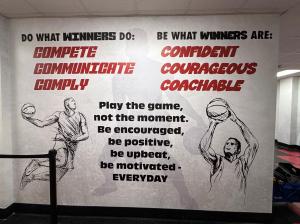 Large lobby wall wrap with sketched basketball players and text including “Compete,” “Communicate,” “Comply,” and “Play the game, not the moment.”