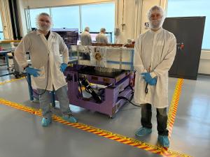 Two people dressed in clean room gear standing in front of a payload for a launch