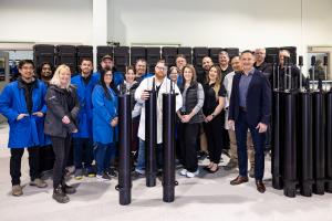 Group of MetOcean Telematics staff standing in a production facility beside multiple black cylindrical NiKA devices, with rows of shipping cases in the background, marking a major shipment milestone.
