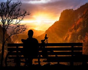 Person sitting on an outdoor bench beside a bicycle, watching the sunset in a calm and reflective setting.