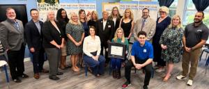 Group of people posing indoors, one seated person holding a framed certificate, others standing around in business-casual attire.