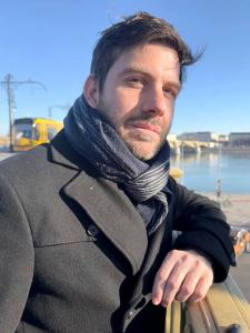 Portrait of B.P. Lemon wearing a black coat and scarf, leaning on a railing by a river with a bridge and city buildings in the background under a clear blue sky.