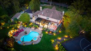 Aerial view of a backyard with a covered deck structure, outdoor dining area, built-in fireplace with TV, pool, patio seating, and landscaped grounds behind a residential home