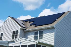 A daytime photo of a two-story gray house. A geometric arrangement of dark solar panels covers most of the brown shingle roof, contrasting sharply with the light siding and a bright blue sky filled with puffy clouds.