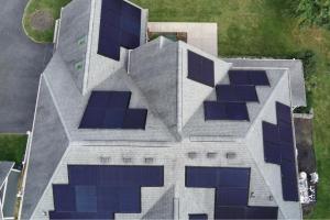 An aerial drone photograph looking straight down on a grey shingle rooftop covered in a patchwork grid of dozens of dark-coloured solar panels, showcasing the intricate system design on a multi-faceted roof.