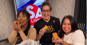 Three people smile on the set of SA Live while holding lumpia. From left to right are host Jada Pickett, Lumpia Week organizer Gene Carangal, and Lumpia Week organizer Nicolette Ardiente, standing in front of an SA Live sign.