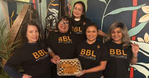 Five members of the Philippine American Chamber of Commerce–Greater San Antonio stand together indoors, smiling and wearing black BBL (Body By Lumpia) shirts, with a tray of lumpia displayed in front of them during Lumpia Week.
