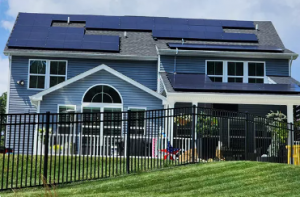 A wide view of a blue-gray residential home in Abingdon featuring an extensive rooftop solar panel array, viewed through a black security fence on a grassy hill.