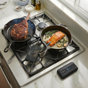 Two pans cooking steak and salmon on a stovetop with MeatStick probes inserted, showing dual-probe monitoring via mobile app and Smart Base display.