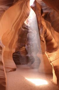 A bright beam of sunlight shining into a narrow slot canyon, lighting up smooth orange sandstone walls and sandy ground.
