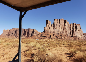 Tall sandstone buttes in Monument Valley viewed from a shaded structure with desert vegetation and open sky.