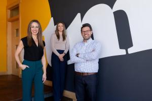 Three coworkers stand smiling in an office in front of a bold black, white and yellow wall mural. A woman in a black top and teal pants stands in the foreground, with a woman in a beige top and navy pants and a man in a checkered shirt standing behind her
