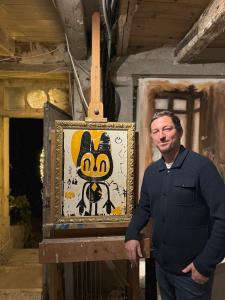 A male artist, smiling, leaning against a painting easel with a contemporary painting of a stylized animal figure in a gold frame.