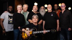 Todd sits in center with guitar, artists line up behind him in dark studio.