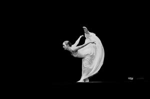 A black-and-white image of a ballet dancer performing a high leg extension on stage. She is balanced on one foot, with her other leg lifted high behind her, arms extended outward. She wears a flowing white dress that creates a soft, sculptural shape again