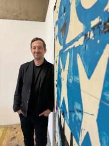 A man in a suit jacket leaning against a wall with a large contemporary abstract painting the foreground, in cream, blue and black.