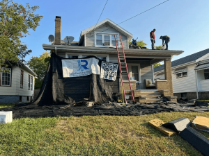 Rainforcing Roofing experts replacing shingles on a residential home. The site is protected with debris netting featuring the company logo and a QR code