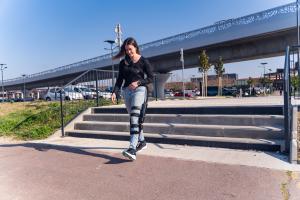 A woman wearing a low profile, robotic KAFO, walking down and away from steps. Background is a parking lot and overpass.