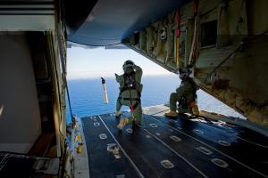 Two U.S. Coast Guard crew members deploy an iSLDMB from the rear of an aircraft over open ocean, releasing the buoy mid-air to track surface drift conditions during a search and rescue mission.