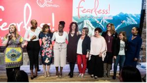 A diverse group of women stand together on a stage at the Fearless Women’s Summit in Toronto, smiling and linking arms in front of a pink and mountain-themed backdrop. One Woman Founder, Sharla Brown, speaks into a microphone while the others face the aud