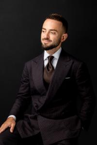 Professional studio portrait of a well-groomed man in a dark textured suit and tie, seated against a black background, looking slightly to the side with a confident expression.
