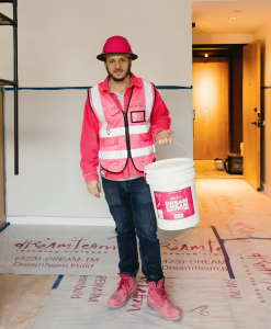 Alejandro Caraballo, Founder of Dream Team Finishes, a Chattanooga, Tennessee commercial finishing company, standing on-site at Caption by Hyatt Chattanooga wearing a pink hard hat and safety vest while holding a bucket of Dream Cream, his proprietary com